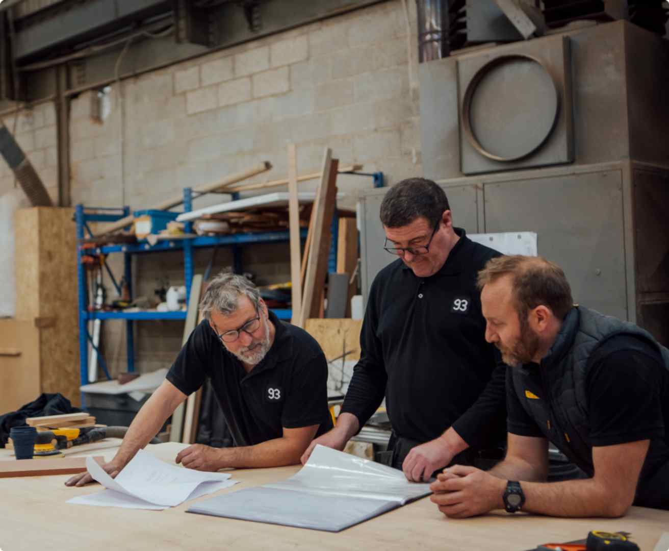 Three men in black polo shirts looking at paper plans in 93 Makes bespoke furniture 