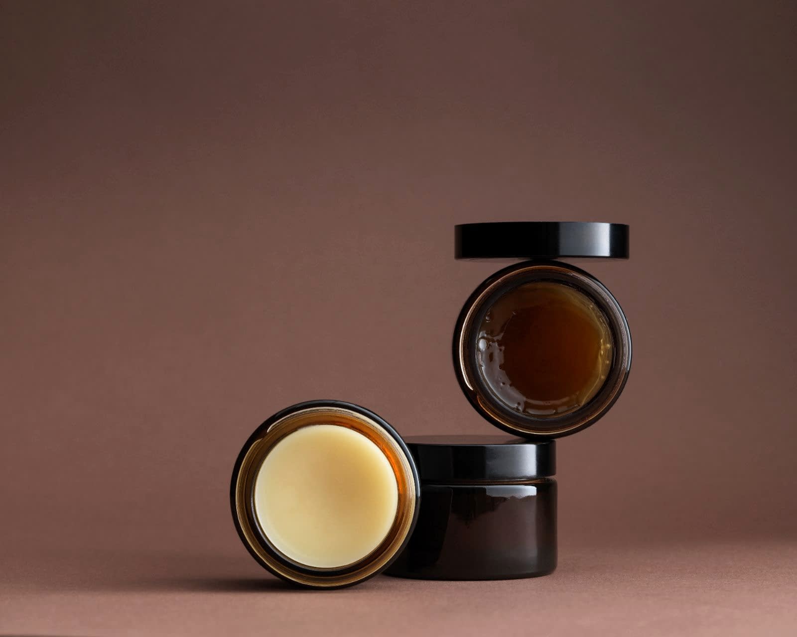 3 brown glass pots stacked artistically with contents facing forward - shiny amber gel and cream pomade. Lid balanced on top.