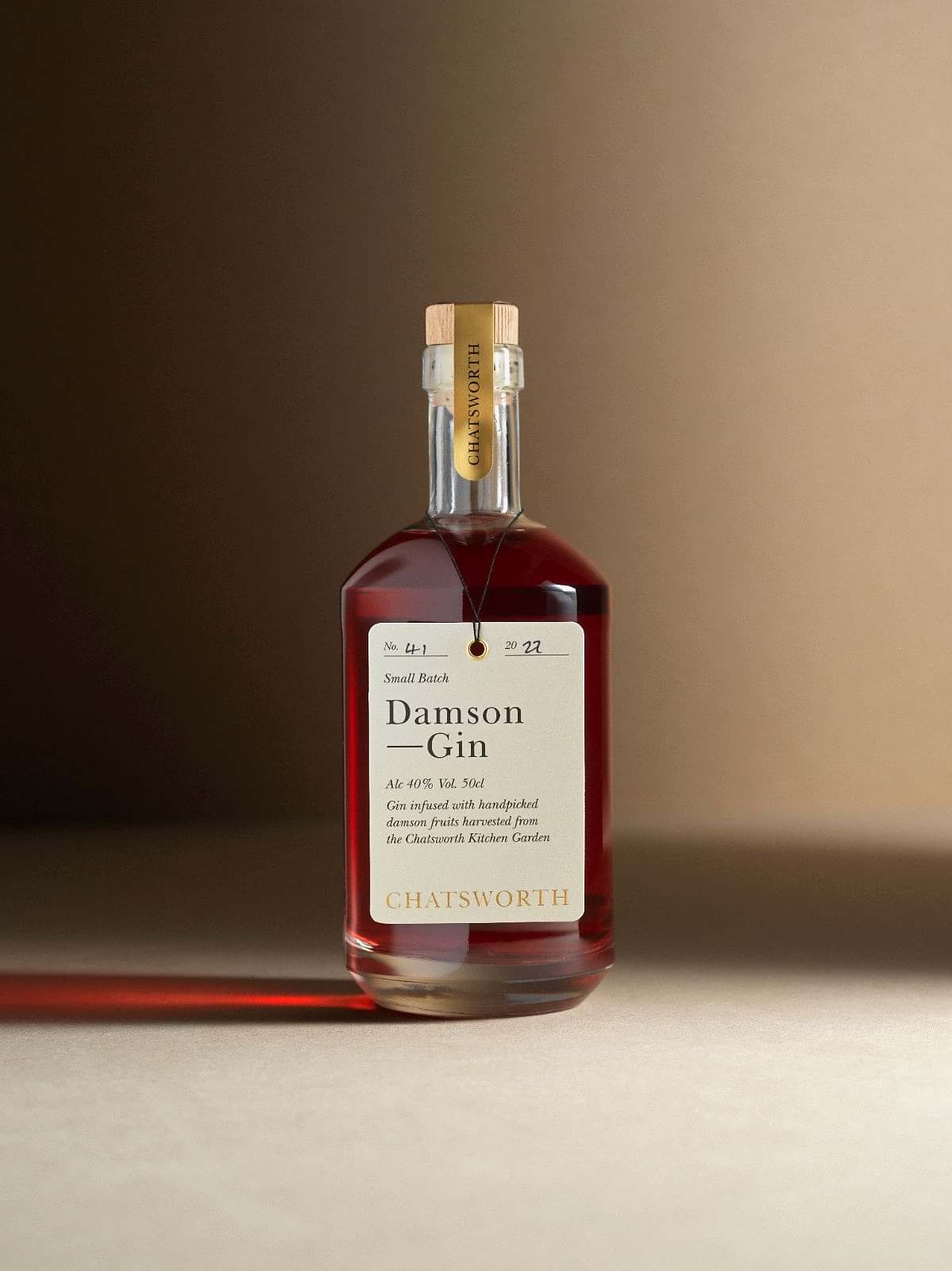 A bottle of red small batch Chatsworth Damson Gin. Clear glass with a cork stopper, gold sticker and cream label.