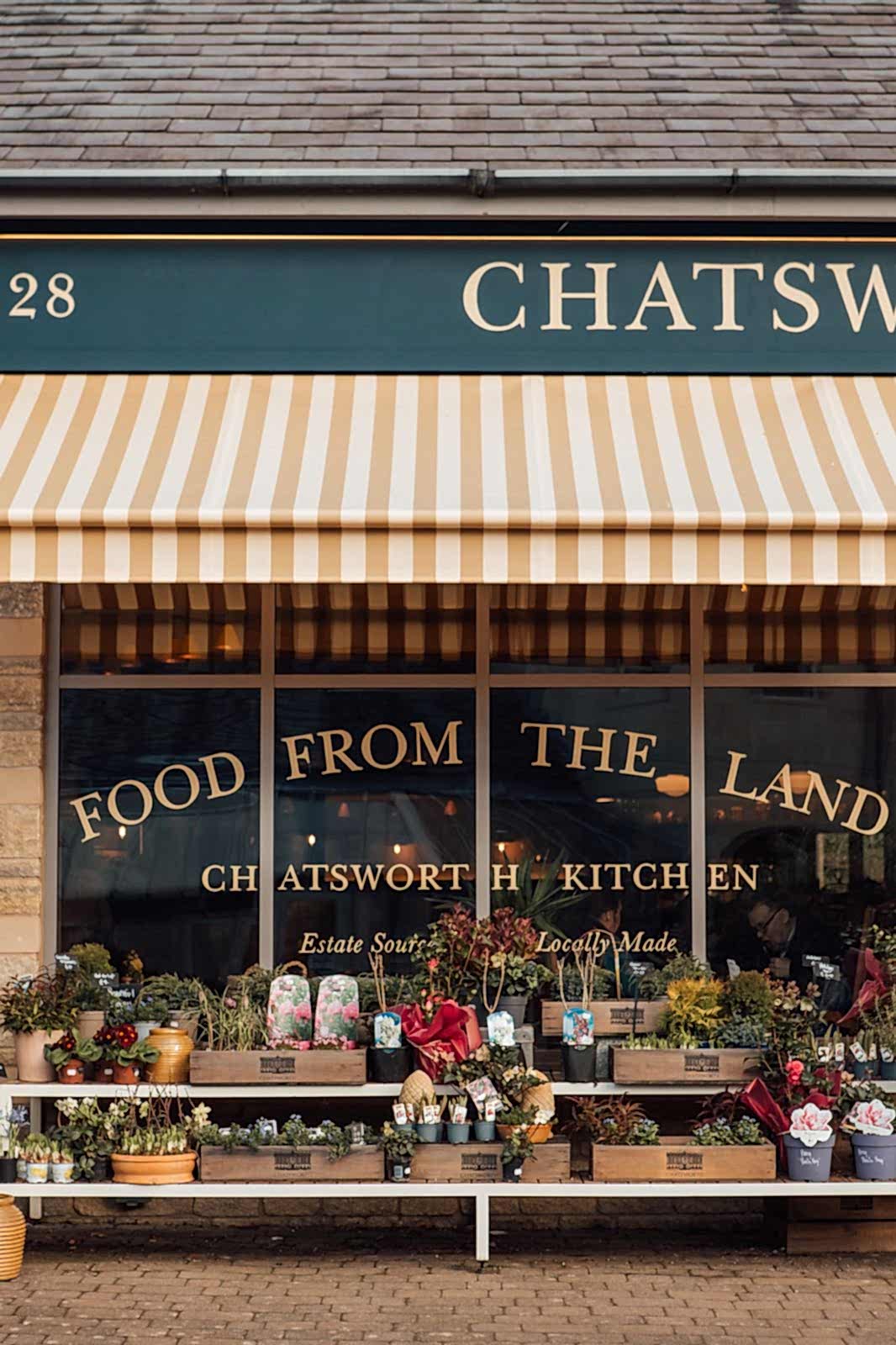 Chatsworth Kitchen facia. Sign writing on windows FOOD FROM THE LAND. Pots of flowers and shrubs outside the building.