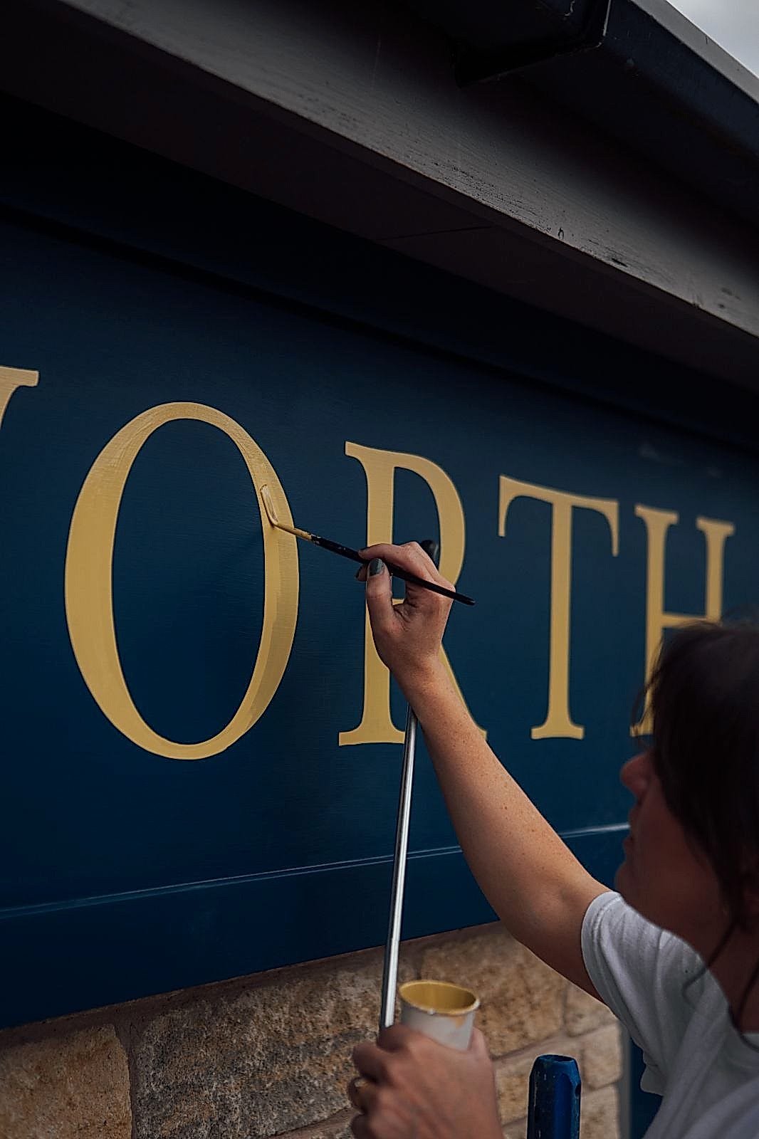 Signwriter Mia Warner painting a gold O on the blue facia sign of Chatsworth Kitchenâ€™s sign at Peak Village. 