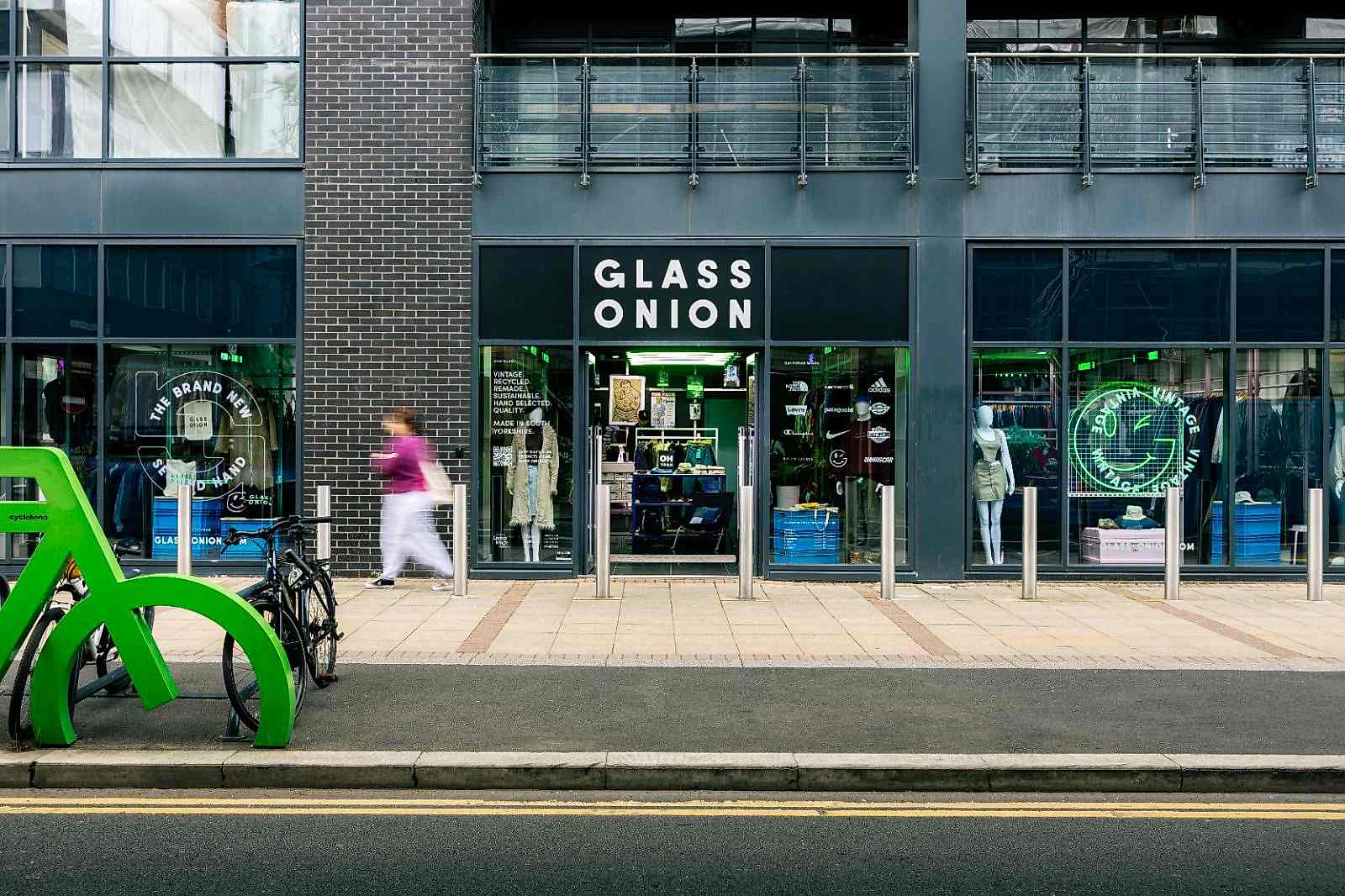 Outside of Glass Onionâ€™s clothing store with a green winking smiley face light in window. Green bike rack in foreground