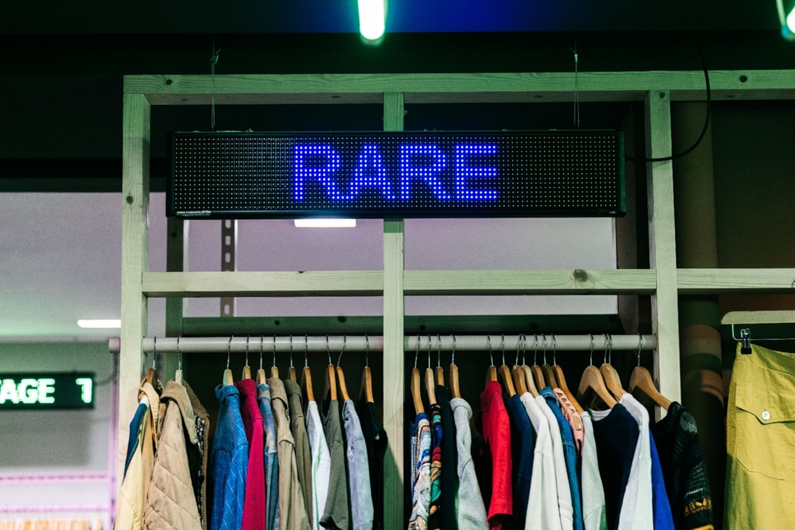 Digital L.E.D signage spelling Rare in purple above a rail of vintage clothing hung from a plain timber frame