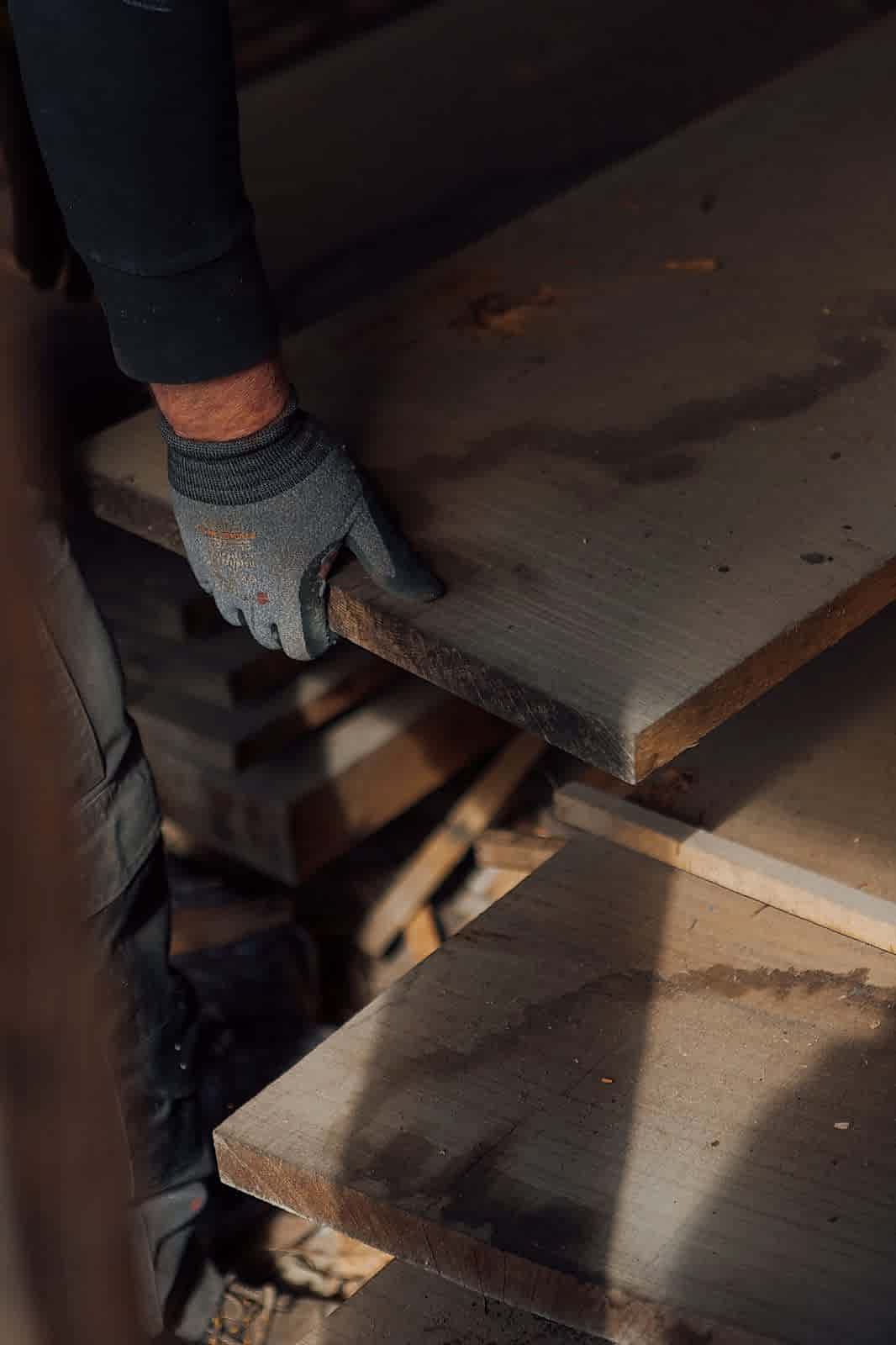 Hand in a work glove picking up a wide plank of reclaimed wood from a stack.