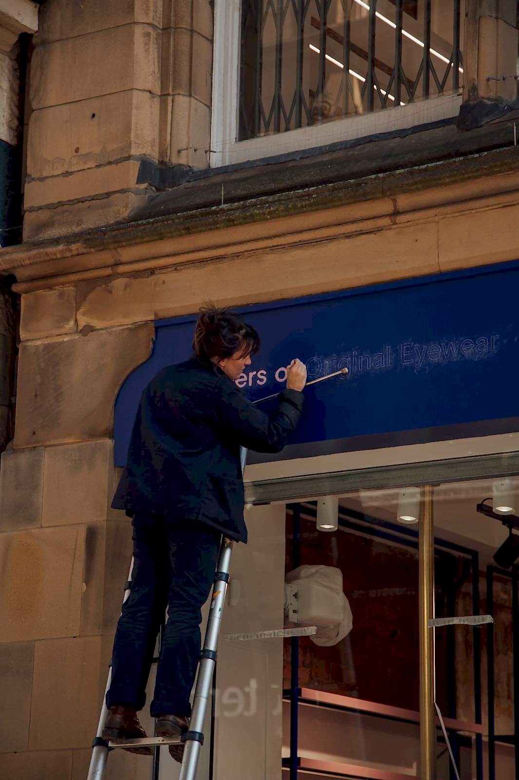 Corner of building with small overhanging pink neon sign of a pair of glasses on blue backing 