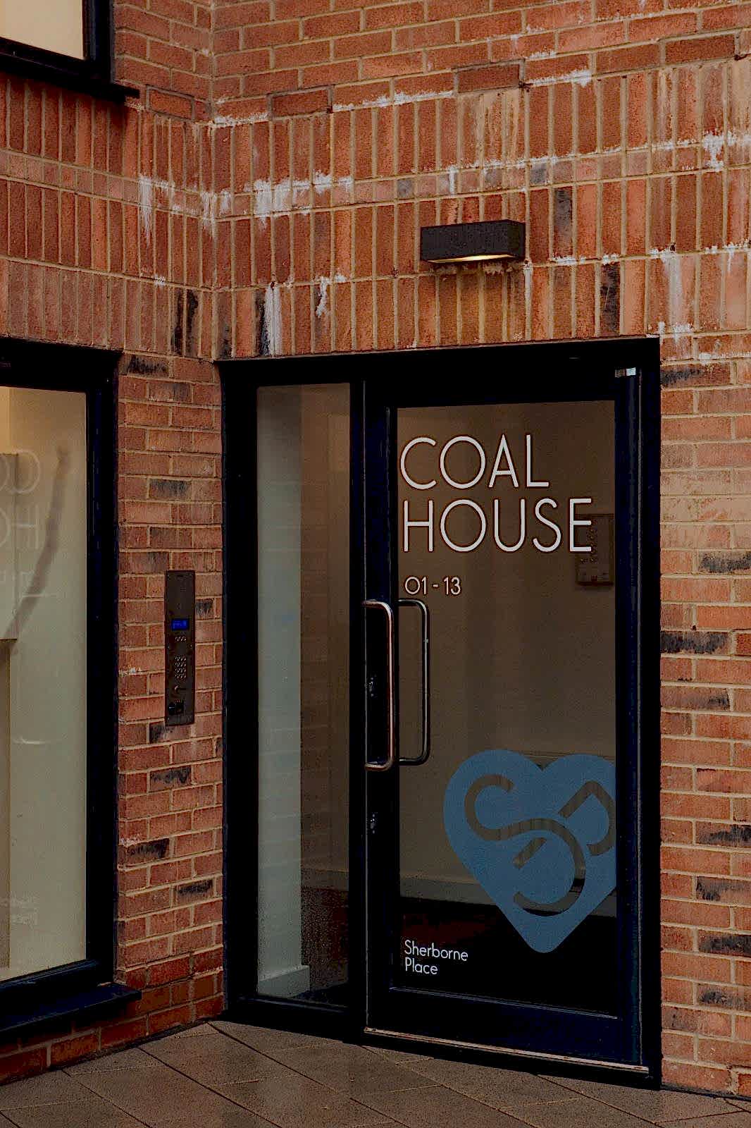 New red brick building with black-framed glass door. Painted on the glass is COAL HOUSE 01-13 and a heart logo with an S.P.