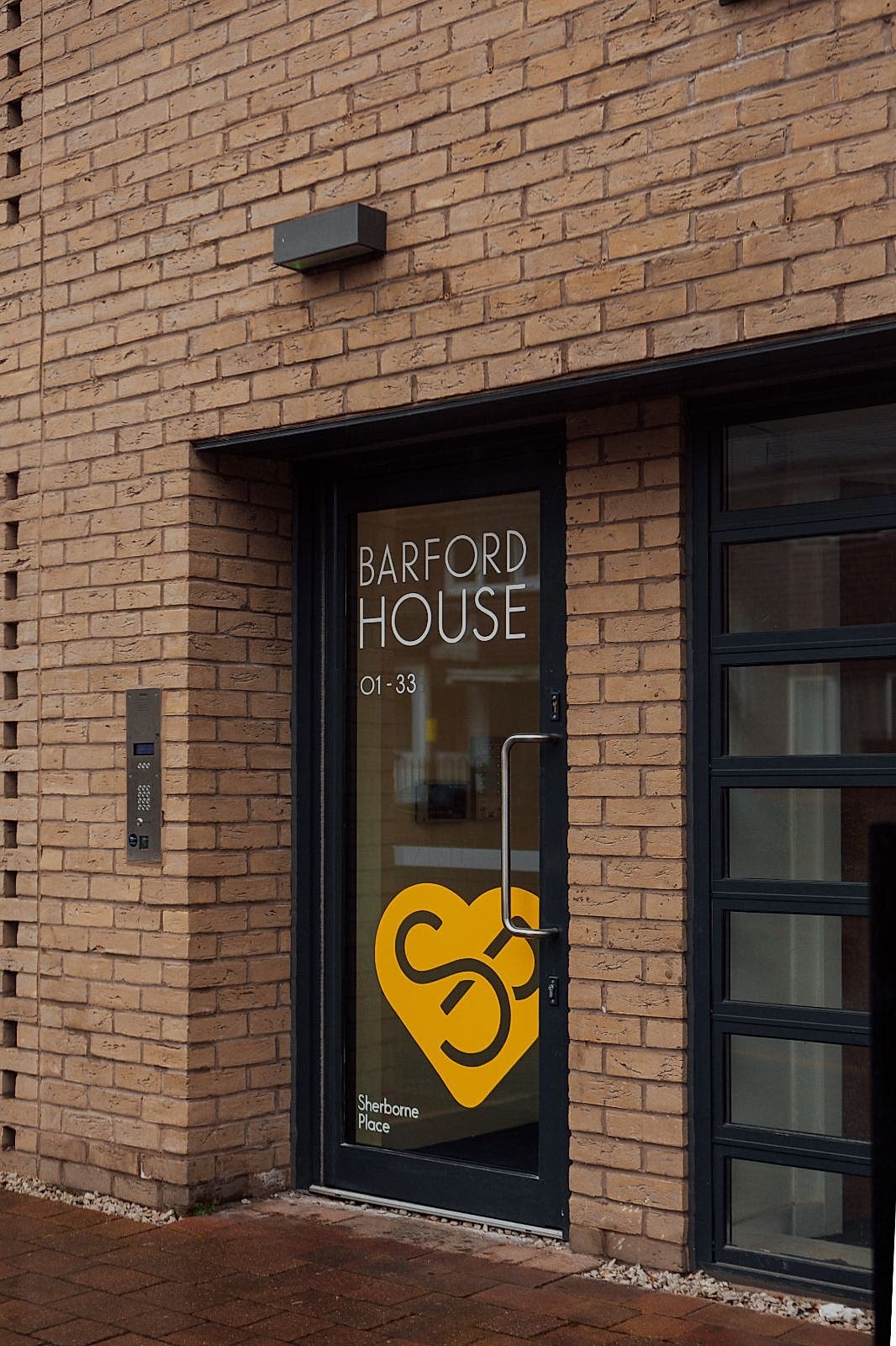 Brick apartment building with black framed door and a window. On the doorâ€™s glass white letters read BARFORD HOUSE