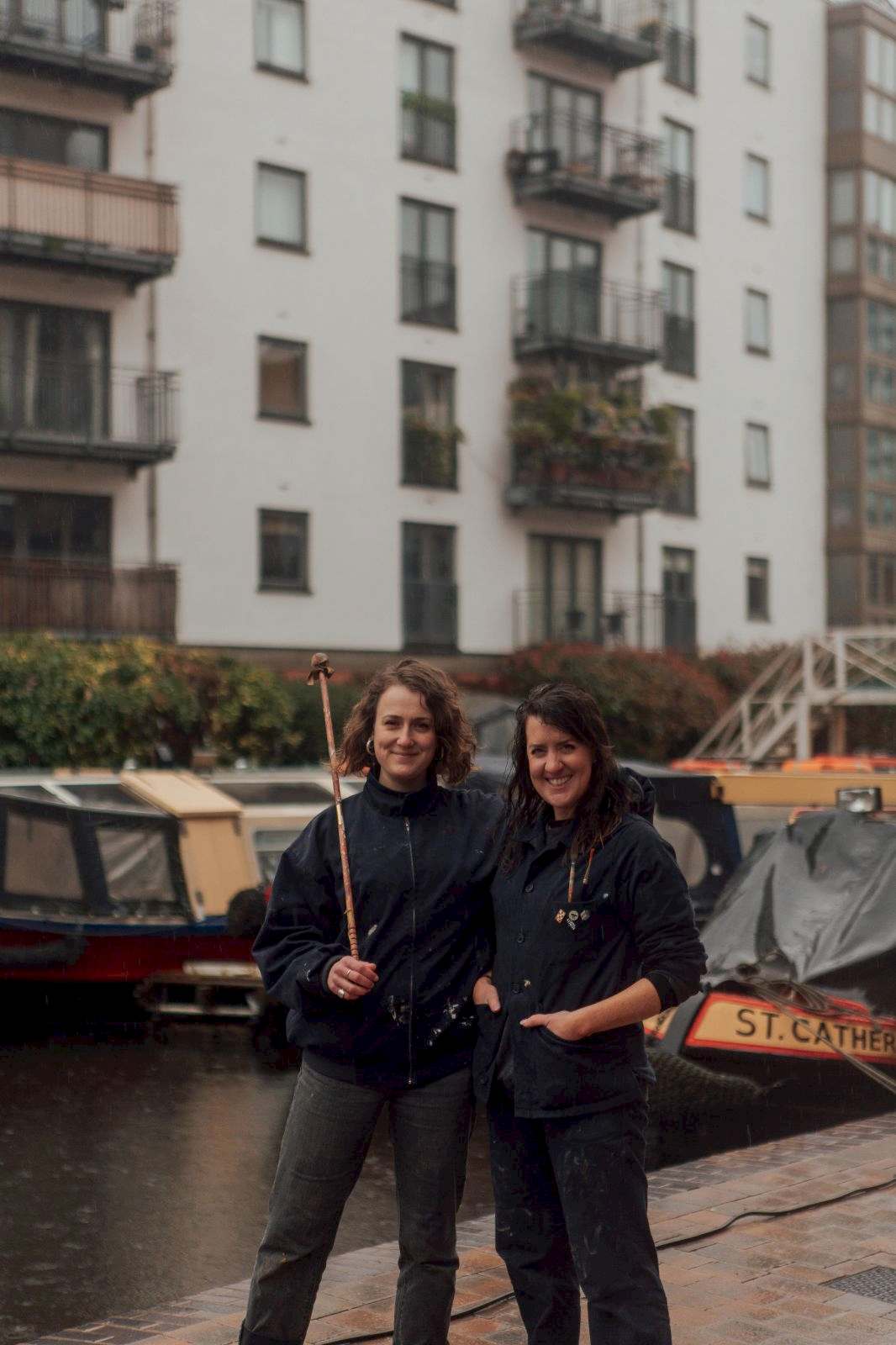 Two female signwriters in dark work clothing, one holds a mahl stick. They sand in front of a wharf with narrow boats.