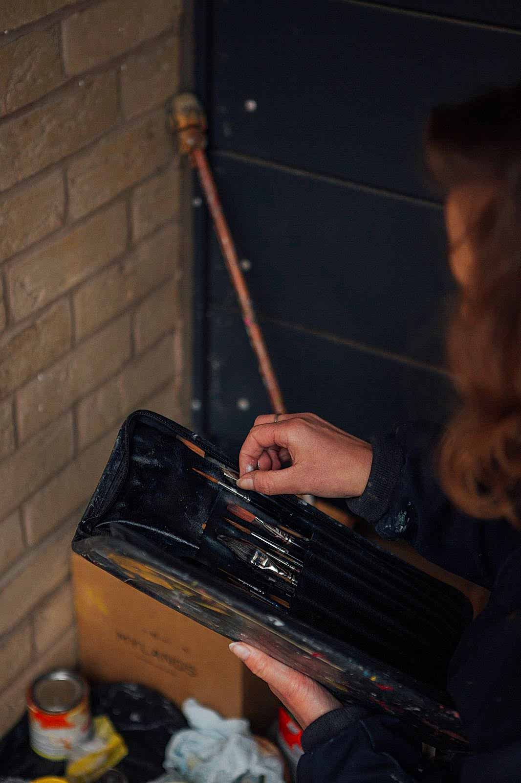 Signwriter reaching into paintbrush case to select paint brush. Below paint cans and bags.