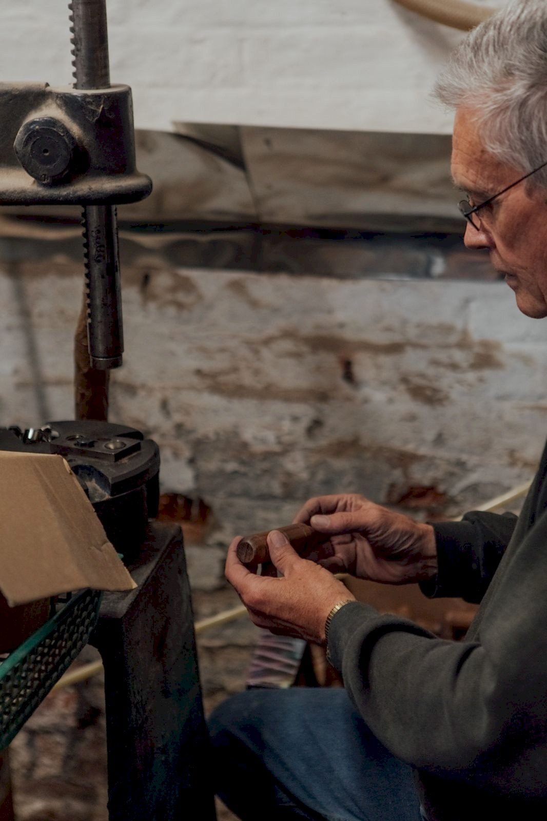 Craftsman holding a turned wooden tool handle in front of a jig. The workshop has old brick walls with peeling paint. 