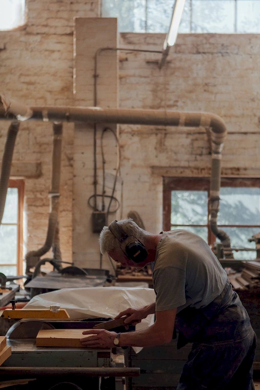 Craftsman cutting wood on a benchsaw inside Woodware Repetitions Workshop. Dust extraction ducts in the background. 