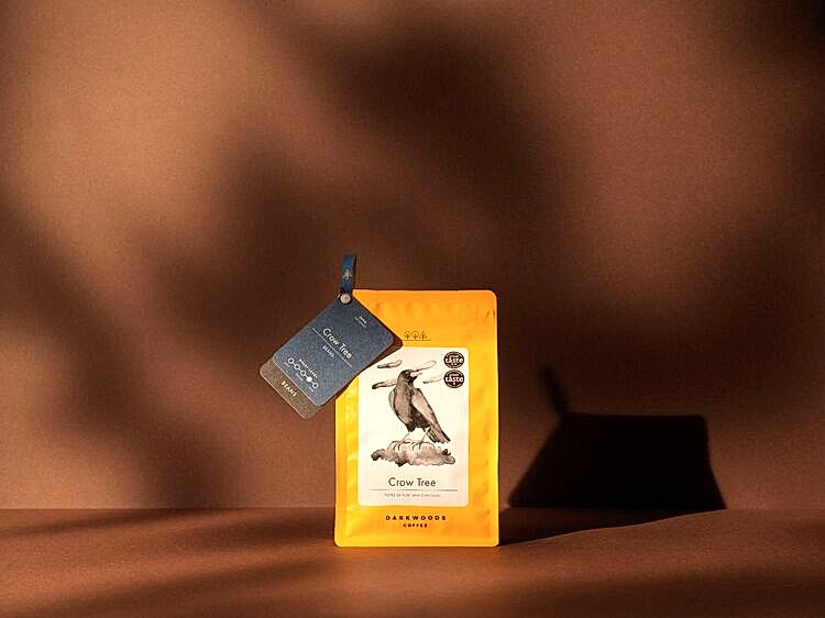 Darkwoods coffee - Yellow Crow Tree packaging, with branding by 93 on dark brown background - warm studio lighting