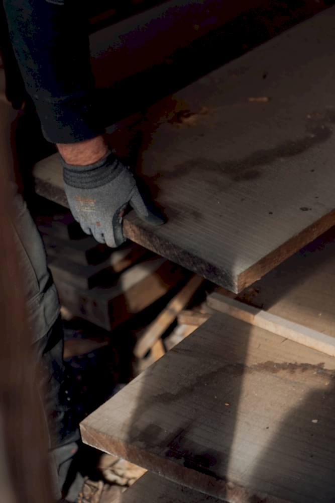 Hand in a work glove picking up a wide plank of reclaimed wood from a stack.