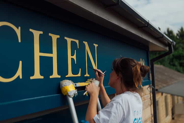 Signwriter painting a black outline around the gold letters of Kitchen on the dark blue shop front facia. 