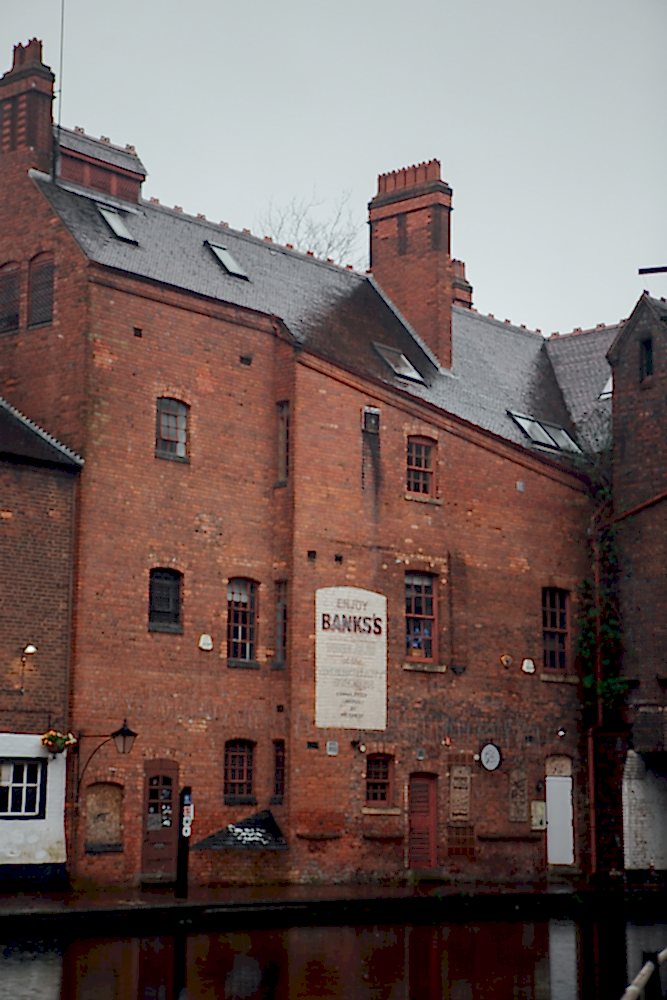 Tall Victorian red-brick building, with painted sign advertising ales, along a Birmingham canal on a rainy winter’s day.