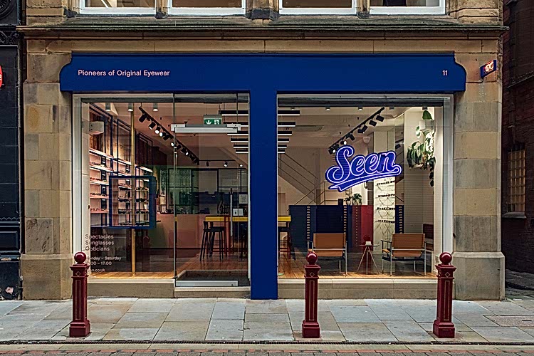 Store front with blue sign facia & large windows. Blue glass neon sign of the Seen logo hangs in the right window.