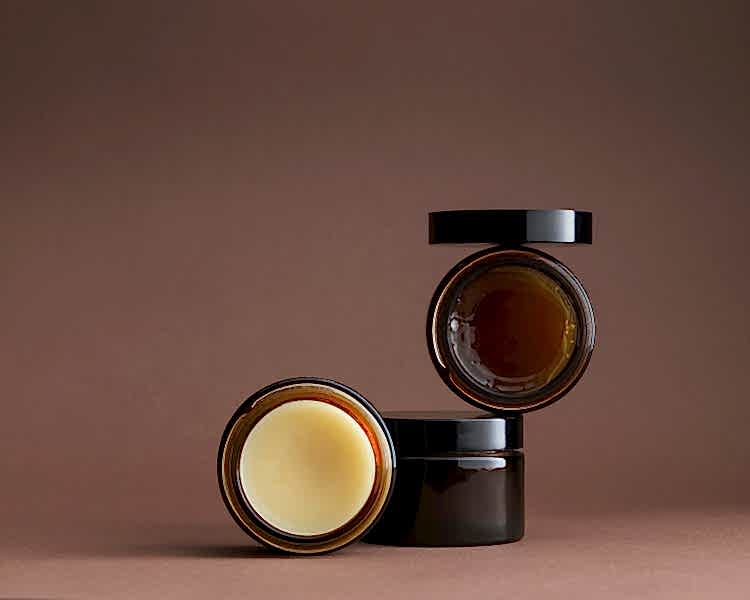 3 brown glass pots stacked artistically with contents facing forward - shiny amber gel and cream pomade. Lid balanced on top.