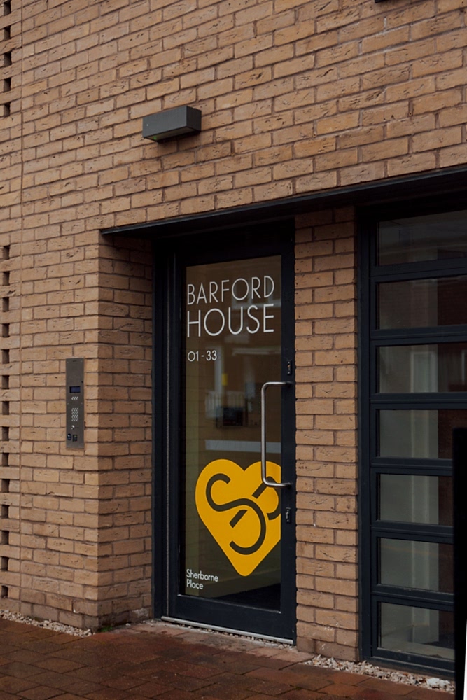 Brick apartment building with black framed door and a window. On the door’s glass white letters read BARFORD HOUSE