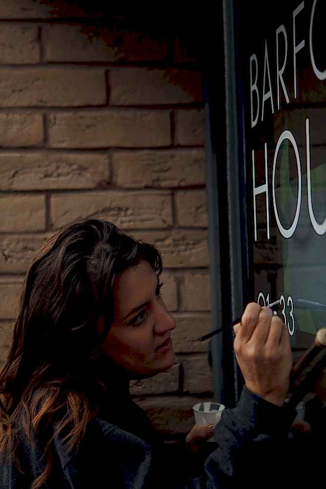 Female signwriter with brown hair painting 33 in white on a glass door. She tilts her head slightly and hand rests on a mahl. 