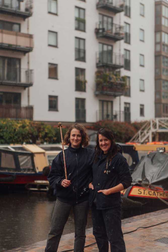 Two female signwriters in dark work clothing, one holds a mahl stick. They sand in front of a wharf with narrow boats.