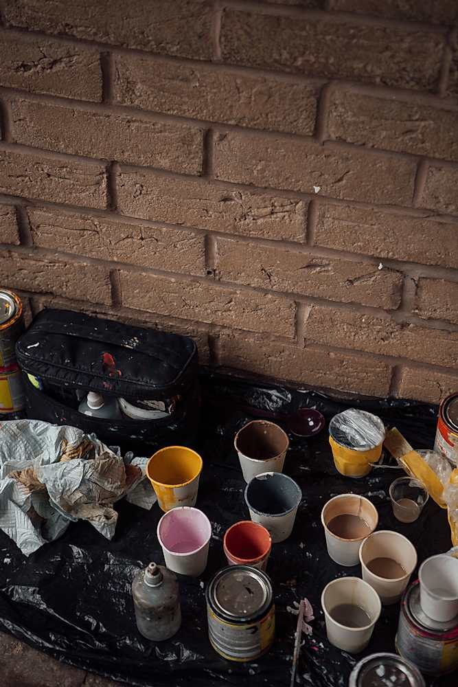 Paper cups of paint in different colours, rags and paint tins on black plastic sheeting on the ground along a brick wall. 