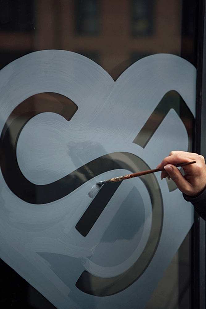 A hand holds a long paintbrush and skillfully paints the S.P heart logo of Sherborne House in blue-grey on glass. 