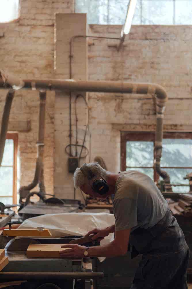 Craftsman cutting wood on a benchsaw inside Woodware Repetitions Workshop. Dust extraction ducts in the background. 