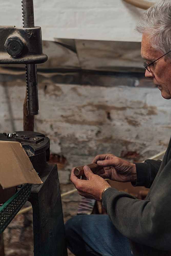 Craftsman holding a turned wooden tool handle in front of a jig. The workshop has old brick walls with peeling paint. 